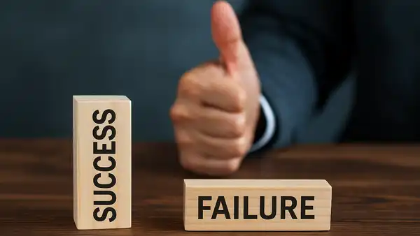Wooden blocks labeled “SUCCESS” and “FAILURE” on a table, with a person giving a thumbs-up in the background